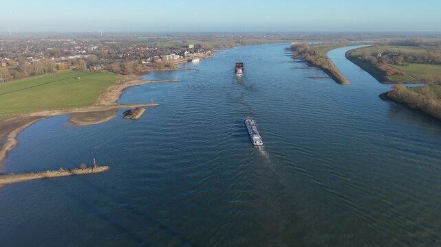 Aerial view of a wide river with two cargo ships moving along calm water, bordered by grassy fields, small settlements, tree patches, and sandbars under a clear sky.