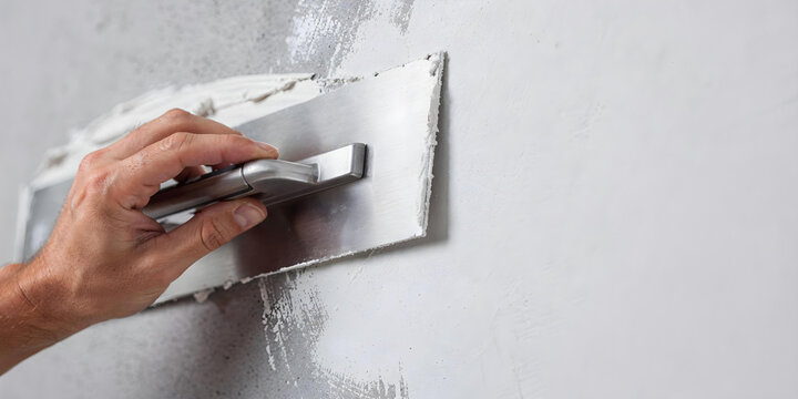 Man hand holding a trowel tool applying white cement plaster or skim coating onto a wall surface. Home improvement and renovation concept.