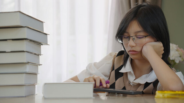 Close-up of a tired female student leaning on her hand, facing a stack of books and a screen, symbolizing modern study stress