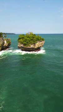 Drone shot showing a dramatic rock island formation rising from the ocean at Karang Bokor Beach, Sawarna, Indonesia. Surrounded by clear sea water and gentle waves in a clean Vertical aerial frame.
