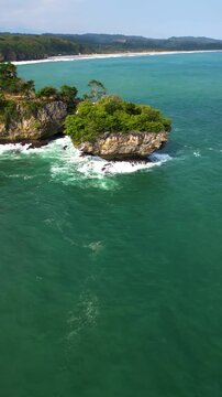 Vertical Aerial drone footage capturing a small rocky islet covered with green vegetation surrounded by clear turquoise ocean water at Karang Bokor Beach, Sawarna, Banten. Natural coastal landscape.