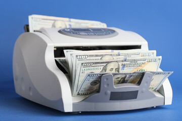 Modern cash counting machine with dollar banknotes on blue background, closeup
