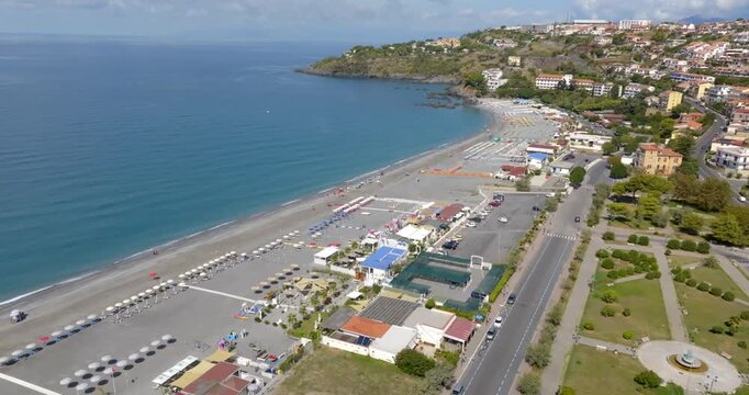 Aerial panorama of a Mediterranean coastline, featuring a vast pebble beach with organized lidos and parking lots, leading toward a rocky promontory. It is the beachfront of Scalea, in Calabria, Italy