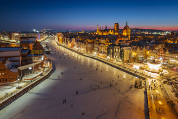 Fototapeta premium Aerial view of Gdansk Old Town at night in winter. Frozen Motlawa river, illuminated historic architecture, pedestrian bridge, and St. Mary's Basilica.