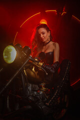 Naklejka na ściany i meble Stylish beautiful young woman in black leather corset and boots posing on vintage motorcycle on old garage dark interior background.