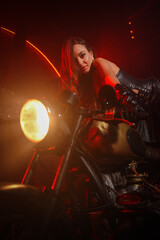Fototapeta na wymiar Stylish beautiful young woman in black leather corset and boots posing on vintage motorcycle on old garage dark interior background.