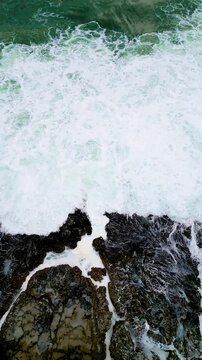 Drone top view of thick sea foam spreading across rugged reef rocks at Karang Bokor Beach, Sawarna, Indonesia. Natural ocean movement and coastal textures recorded in smooth Vertical format.