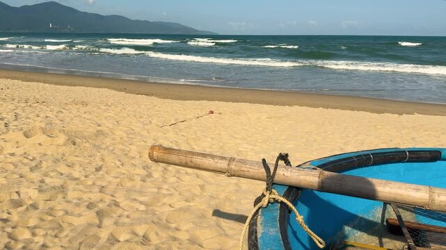 Traditional vietnamese blue basket boat with a wooden paddle resting on the golden sand of my khe beach, with turquoise ocean waves rolling towards the shore and distant mountains under a blue sky
