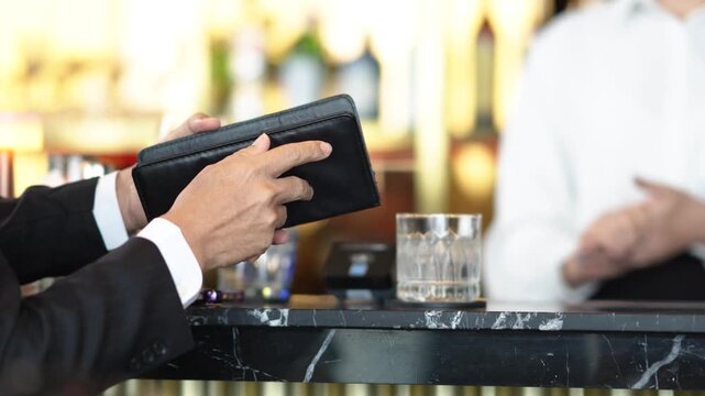 Close up video of businessman hand receiving bill folder at bar counter while bartender prepares drink in background. Corporate nightlife and payment concept.