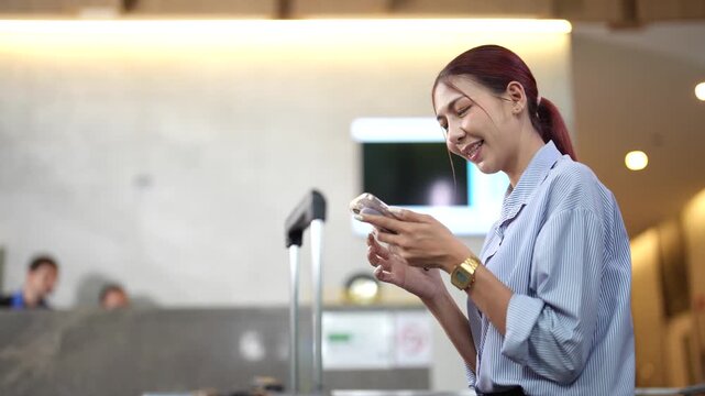 Young Asian woman waiting in hotel lobby and scrolling on smartphone while reception team assists guests behind counter. Travel convenience and connected lifestyle concept.