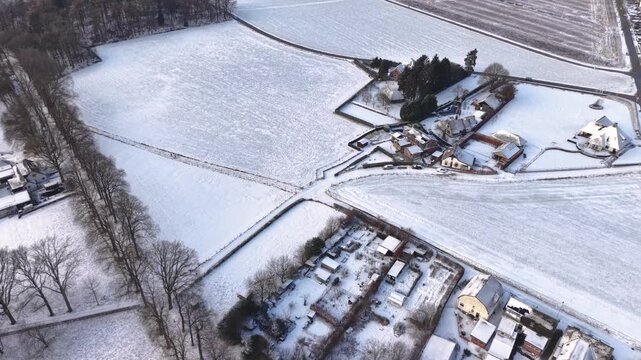 Aerial view of a snow covered rural area with scattered houses, tree lined paths, open fields, and quiet winter roads forming a calm countryside landscape.
