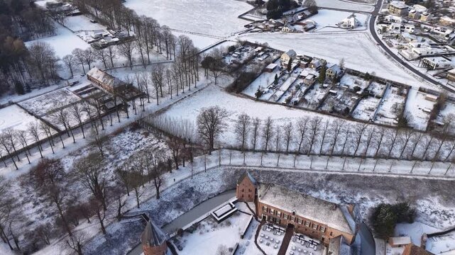 Aerial view of a snow covered rural area with scattered houses, tree lined paths, open fields, and quiet winter roads forming a calm countryside landscape.