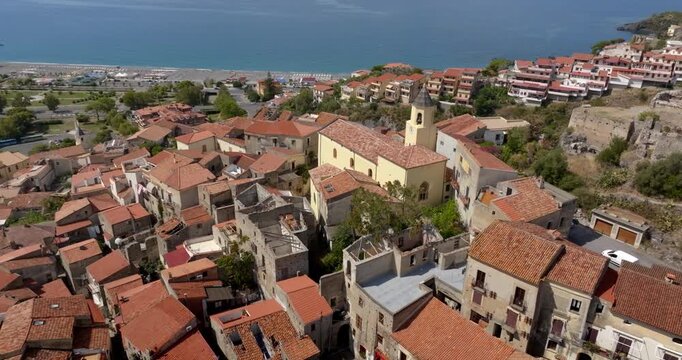Aerial view of an Italian coastal hamlet. Terracotta rooftops and a prominent church tower lead the eye toward a beachfront and the deep blue Mediterranean sea. Panorama of Scalea, in Calabria, Italy.