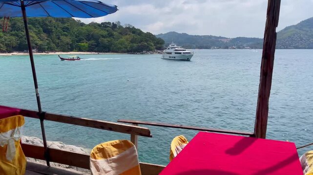 Relaxing view from a seaside restaurant in Phuket, overlooking to yacht on turquoise waters, long tail boat cruising the bay, and lush tropical hills under a bright sky