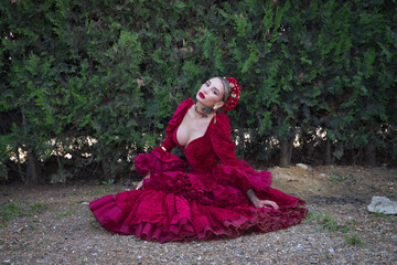 Fototapeta premium Young, beautiful, blonde woman in red flamenco dress, flowers in her hair and gold jewelry, performing flamenco poses sitting on the ground with green plants in the background. Concept dance, art.