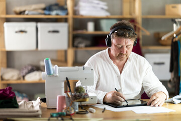 Fototapeta premium Middle-aged male dressmaker wearing earphones drawing on tablet in sewing workshop