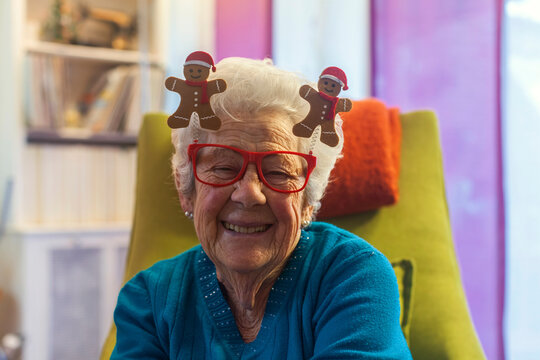 Festive portrait of smiling senior woman wearing Christmas gingerbread glasses and santa hat boppers at home. Cheerful elderly grandmother celebrating the holidays. Authenti