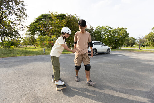 Asian Girl Learning Skateboarding While Holding Brother&rsquo;s Hand Outdoors