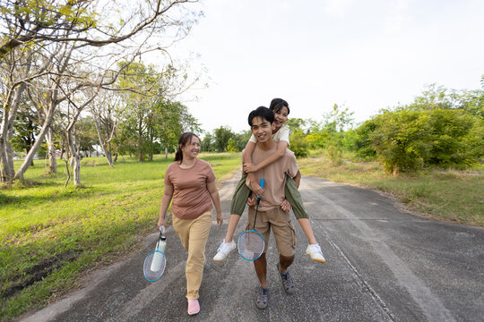Asian Family Walking in Public Park with Brother Carrying Sister and Badminton Rackets