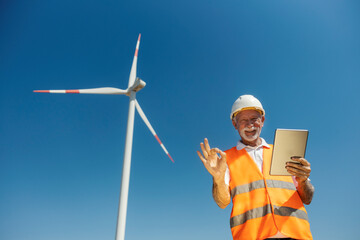 Senior Male Environmentalist Inspecting Windmill Holding Tablet Wearing Safety Gear Outdoors