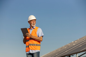 Senior White Male Environmentalist Inspecting Solar Panels with Tablet on Solar Farm Under Clear Sky