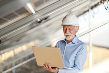 Senior White Male Environmentalist Inspecting Solar Farm Using Laptop Among Solar Panels
