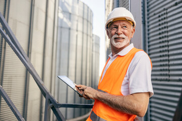 Senior male industrial worker in orange vest using tablet outdoors at factory facility