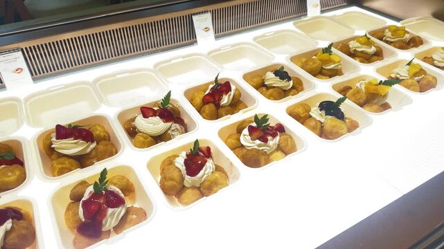 Cream puffs topped with whipped cream and fresh strawberries, mango and blueberries, neatly packed in individual compostable takeout boxes ready for sale at a dessert shop