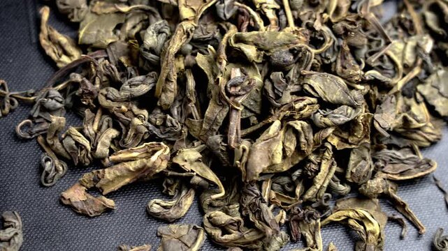 A pile of curled scattered dried green tea leaves on a black cardboard packaging box surface
