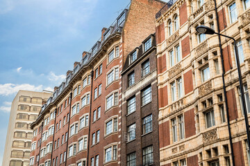 Fototapeta premium Classic British architecture lines a traditional London street, with red brick buildings and historic facades beneath a cloudy sky.