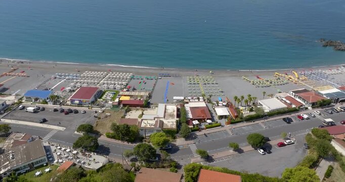 Aerial view of a vibrant coastal resort featuring organized beach umbrellas, turquoise waters, and structured seaside buildings. It's the beachfront of Scalea, in Calabria, southern Italy.