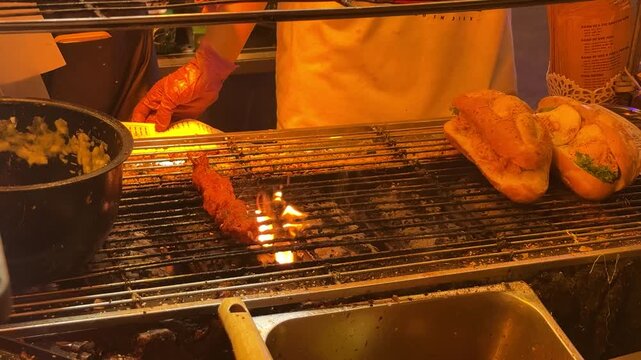 Street vendor's gloved hand turning seasoned meat skewer over hot charcoal grill while toasting banh mi rolls nearby, capturing lively da nang night market cuisine ambiance