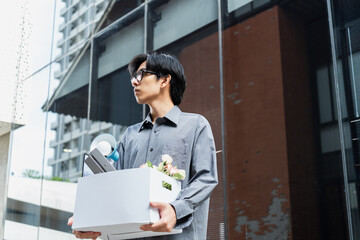 Stressed young man in glasses carrying personal items in a white box after job loss. Concept of unemployment.