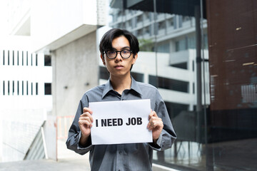 Serious young Asian man holding a sign that says "I NEED JOB" in front of an office building. Concept of unemployment and job searching.