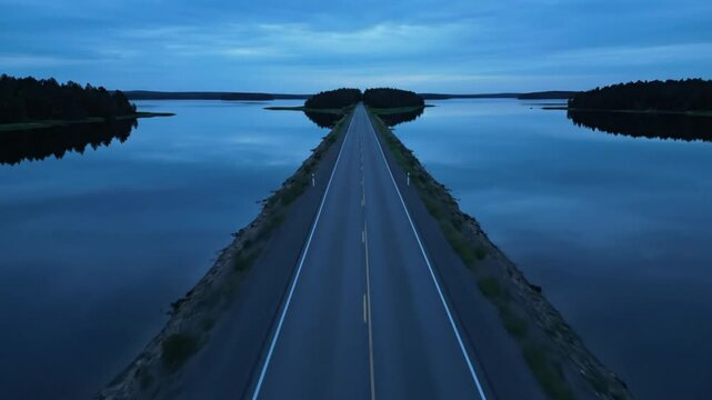 Drone perspective of a long straight road running through calm water at twilight with reflecting trees and sky