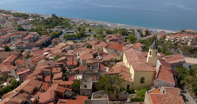 Aerial view of an Italian coastal hamlet. Terracotta rooftops and a prominent church tower lead the eye toward a beachfront and the deep blue Mediterranean sea. Panorama of Scalea, in Calabria, Italy.