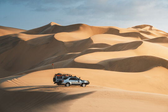 View of a silver SUV navigating through the undulating golden dunes under a serene sky, creating a striking contrast of adventure and tranquility, Walvis Bay, Namibia.