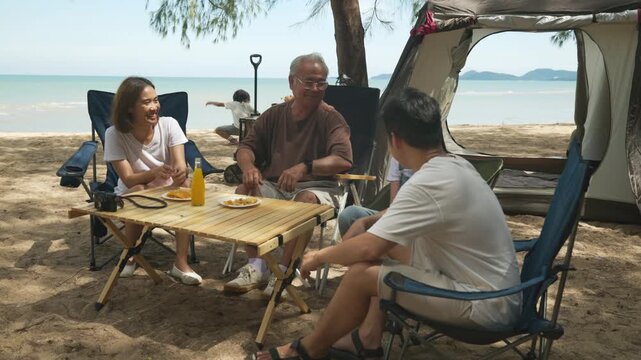 Beach camping family fun trip, kids play ocean side, adults share happy outdoor meal on portable table near spacious camping tent, creating relaxed multi generational vacation memories.