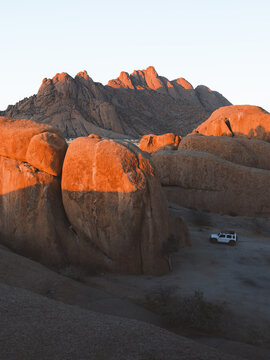 View of warm sunlight paints the rugged rocks, creating a striking contrast with the dark shadows, as a lone vehicle sits in the arid landscape, Erongo, Namibia.