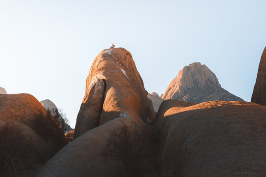 View of massive granite boulders glow in the warm light of the setting sun, a lone figure perched atop, evoking a sense of solitude, Spitzkoppe, Erongo Region, Namibia.