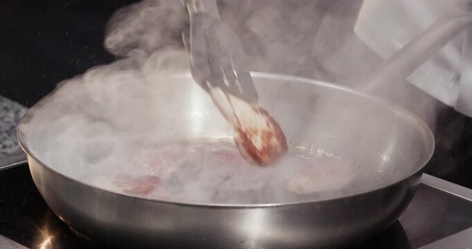 Chef turns beef slices with tongs on hot frying pan in kitchen closeup. Master cooks delicacy dish with veal in fancy restaurant. Healthy dinner preparation