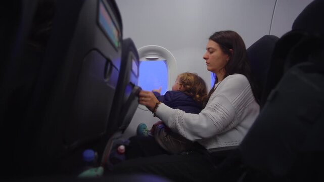 Toddler seated on mother lap opening and closing airplane tray table while exploring cabin evoking curiosity learning and calm preflight engagement
