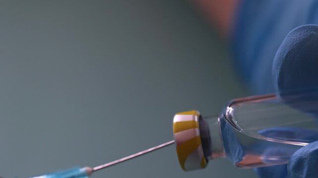 Extreme close-up of a needle piercing a vial stopper while a gloved hand pulls the syringe plunger to draw liquid medication. Medical preparation and vaccination concept.