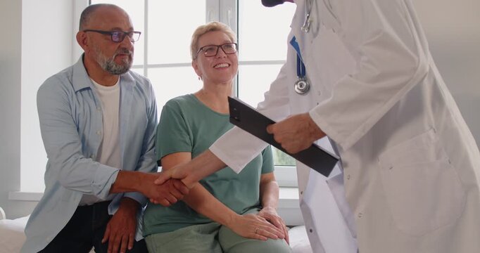Doctor offers a warm handshake to seniors, welcoming patients and couple before an appointment in the clinic. Physician greets mature family in medical office during exam. Medicine concept.