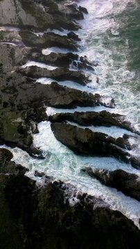 Vertical Drone footage capturing dramatic layered rock formations with ocean water flowing between the gaps at Karang Taraje Beach, Sawarna, Indonesia. Strong coastal textures and white foam movement.