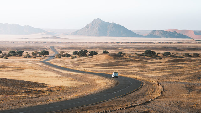 View of a lone white car winding its way through the arid landscape under a hazy sky, with distant mountains rising, reflecting the sun's glow, Windhoek, Hardap Region, Namibia.