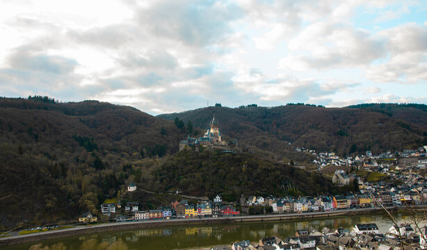 View of a medieval castle perched atop a hill, overlooking a colorful town reflected in the river below, under a cloudy sky, Cochem, Rheinland-Pfalz, Germany.