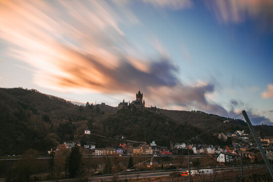 View of the imposing castle perched atop a tree-covered hill, overlooking the quaint town nestled below as clouds streak across the sky, Cochem, Rheinland-Pfalz, Germany.