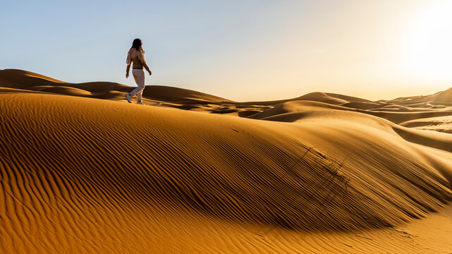 A woman walks across a vast, undulating sand dune landscape under a clear sky. Mezouga,Sahara desert,Morocco