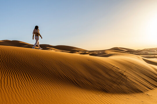 A woman walks across a large sand dune, with more dunes in the background under a clear sky. Mezouga,Sahara desert,Morocco